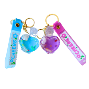 A pair of floating heart liquid keychains with gold-tone keyrings, featuring a cute bear inside a transparent heart filled with glittery liquid and floral confetti, attached to a "CHEERS" printed silicone strap in blue and pink variations.