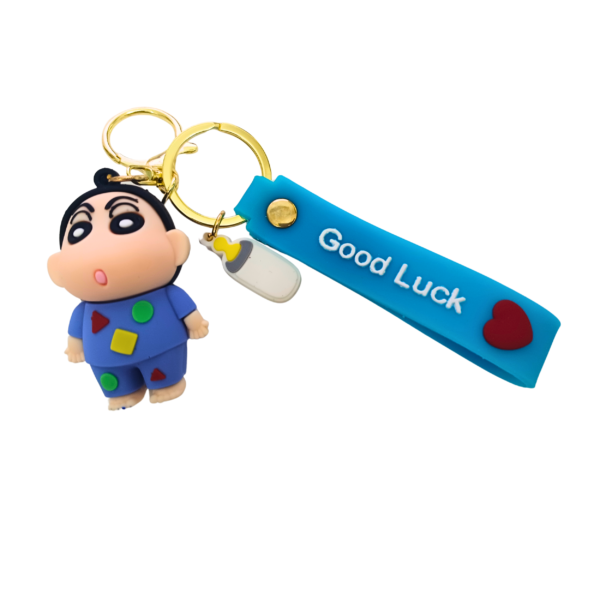 A Shinchan keychain featuring him in a blue geometric-patterned outfit, attached to a blue "Good Luck" silicone strap and a gold keyring.